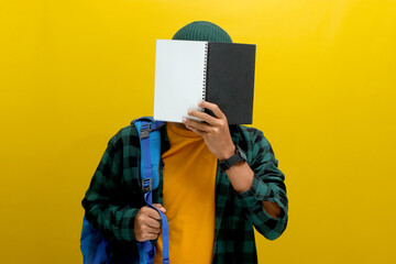 A young Asian man, dressed in a beanie hat and casual shirt and carrying a backpack, is covering...