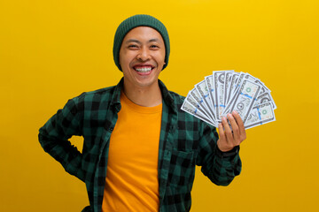 Excited Asian man in a beanie and casual clothes holds banknotes in his hand. Isolated on a bright...