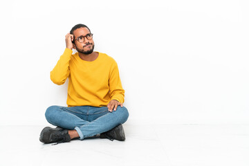 Young Ecuadorian man sitting on the floor isolated on white wall having doubts and with confuse...