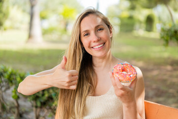 Young blonde woman holding a donut at outdoors with thumbs up because something good has happened