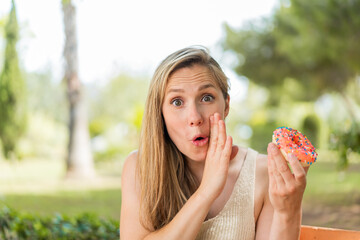 Young blonde woman holding a donut at outdoors whispering something