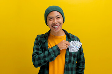 Cheerful Asian man in a beanie and casual clothes points to overflowing pockets full of cash,...
