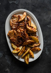 Delicious lunch, dinner - pork chop, baked new potatoes and caramelized onions in one plate on a dark background, top view