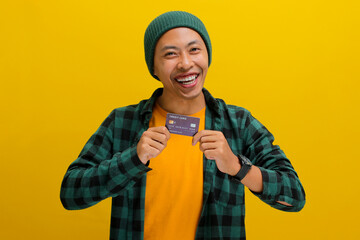 A happy young Asian man, dressed in a beanie hat and casual shirt, enthusiastically shows a credit card in his hand, feeling excited while demonstrating a solution for online shopping payment