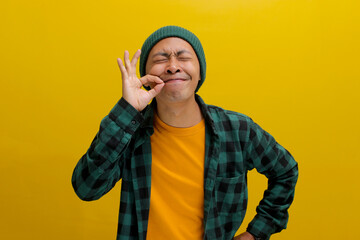 Joyful young Asian man, closes his eyes and uses his fingers to make a gourmet or delicious hand gesture, expressing how enjoyable the food is while standing against a yellow background