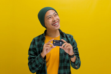 Pensive young Asian man, dressed in a beanie hat and casual shirt, holds a credit card and gazes at...