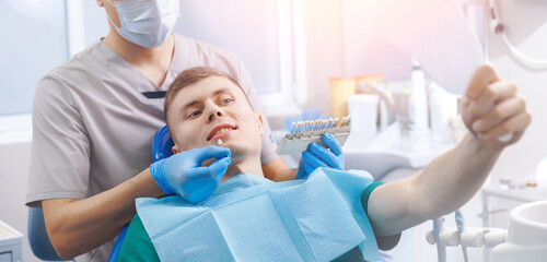 Young man smiling while looking at mirror in dental chair after professional cosmetic whitening...