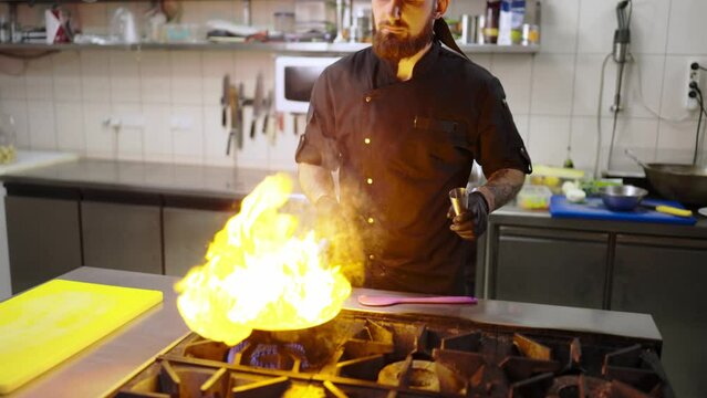 Confident male chef in a black uniform and with black protective gloves pours alcohol onto fried vegetables in a frying pan to apply the flambing technique in a modern kitchen in a restaurant