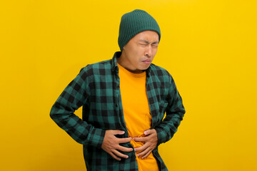 Unhappy Asian man is holding his belly in pain, visibly suffering from a stomachache, likely due to food poisoning, experiencing discomfort, Isolated on yellow background.