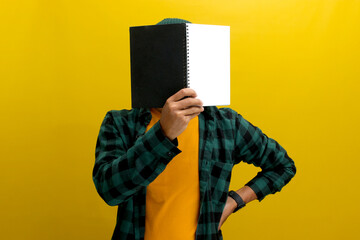 Young Asian man is covering his face with a book while standing against yellow background.