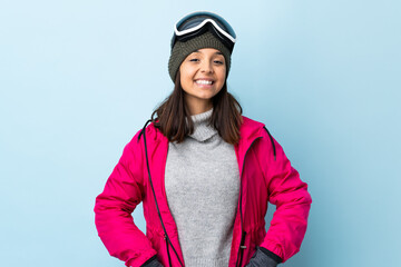 Mixed race skier girl with snowboarding glasses over isolated blue background posing with arms at...