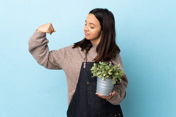 Young brunette mixed race woman holding a plant over isolated blue background doing strong gesture.