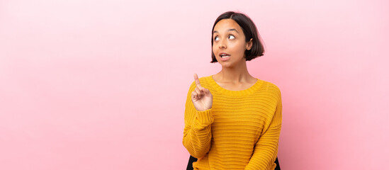 Young mixed race woman sitting on a chair isolated on pink background thinking an idea pointing the...