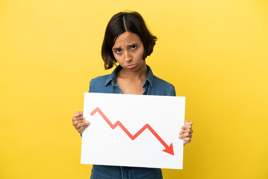 Young mixed race woman isolated on yellow background holding a sign with a decreasing statistics arrow symbol with sad expression