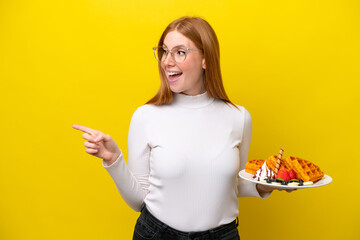 Young redhead woman holding waffles isolated on yellow background pointing finger to the side and presenting a product