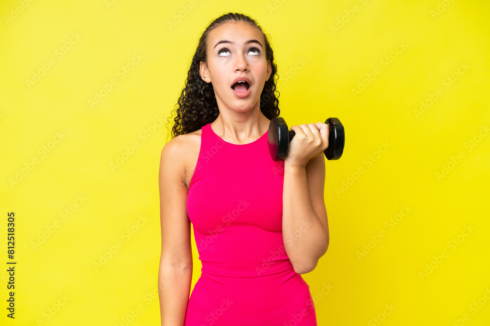 Wall mural Young sport woman making weightlifting isolated on yellow background looking up and with surprised expression