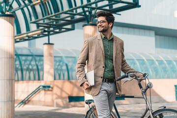 Young cheerful male financial advisor on the way to work with bike holding digital tablet and...