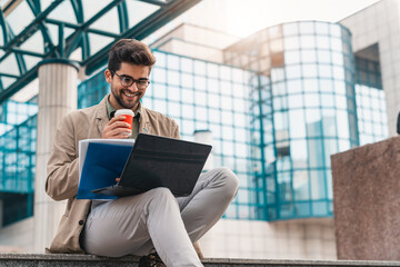 Young happy businessman using laptop outside of modern work place. Smiling man with glasses...