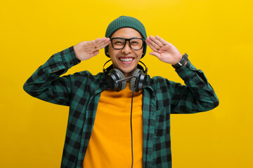 A young Asian man, dressed in a beanie hat and casual clothes, salutes with a cheerful expression and a smile directed at the camera while standing against a yellow background