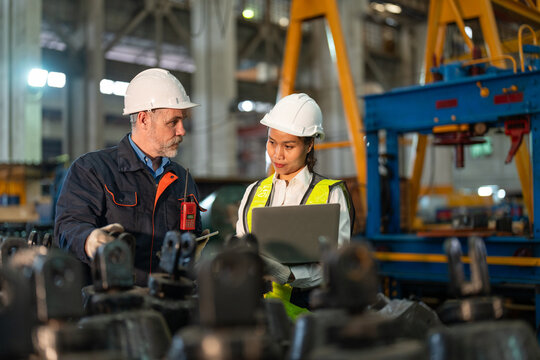 Senior male engineer and Asian female engineer collaborate using a laptop amidst heavy machinery in an industrial factory setting.