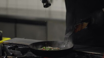 Close up a professional chef in a black uniform pours alcohol onto a hot frying pan with a dish to create fire and flambéing technique while preparing a dish in a restaurant kitchen. The cook adds
