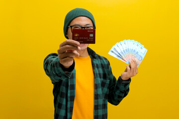 Thrilled Asian man in a beanie and casual clothes holds up a credit card and a stack of banknotes,...