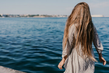 A woman is walking on the beach wearing a dress. The water is calm and the sky is clear.