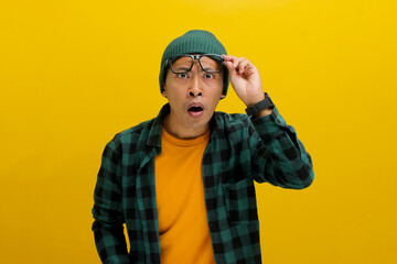 Thrilled Asian man in a beanie and casual shirt adjusts his glasses, eyes wide with surprise as he...