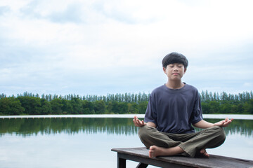 Meditate and relax Asian young boy Smiling and charming sitting and relax at lakeside on natural and sky sunrise background,concept of exercise and healthy,children blue shirt, children sit on floor.