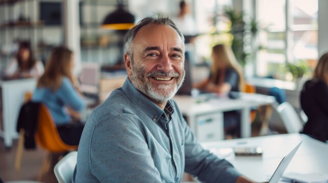 Smiling man with gray beard and hair wearing blue shirt with red pattern sitting at white table in modern brightly lit cafe with other patrons.