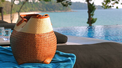 Woven beach bag and sandals resting on lounge chair beside tranquil resort pool overlooking serene tropical beach. Luxury travel and vacation.