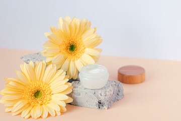 Cosmetic cream jar on stone podium with yellow gerbera flowers on beige table. Closeup