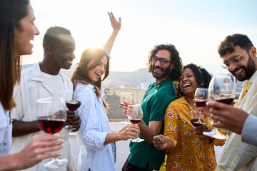 Happy international community enjoying party dance with glass of red wine at sunset rooftop party. Girl dancing with raised hands at disco outdoors with friends. Beautiful people having fun together 