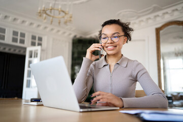 A cheerful young woman engages in a phone conversation while working on a laptop in an elegant, traditional office setting.