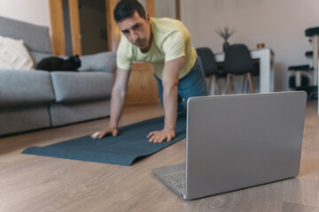 Concentrated man on yoga mat follows online class, stretching at home while focused on laptop...