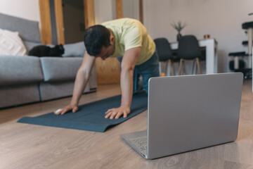 Man Stretching on Mat with Laptop for Online Class in Home Setting. Man does yoga stretches on mat with laptop nearby for online class in cozy home environment.