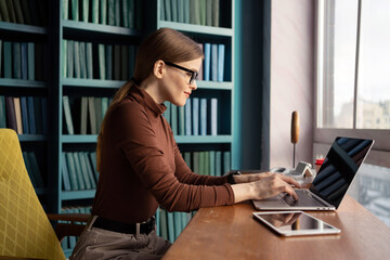 A concentrated young woman works on a laptop in a stylish library setting, surrounded by...