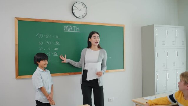 Cheerful student boy writing answer math formula on blackboard with chalk during lesson at school. Young teacher woman and diverse pupils clap hands to classmate correct answer in mathematics.