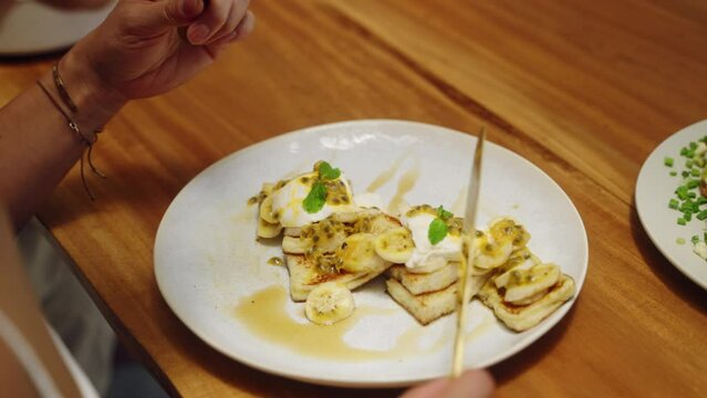 Close-up, hand cuts gourmet banana passion fruit pancakes on wooden table. Person enjoys homemade breakfast, food vlog style. Quick, easy recipe demo for delicious, healthy morning meal.