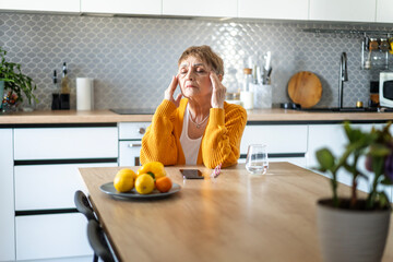 Senior Caucasian woman sitting at the table in the kitchen holding her head, headaches concept