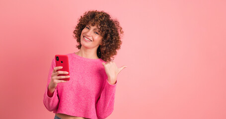 Young woman with mobile phone point to pink wall on pink background