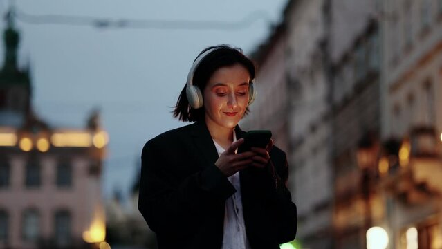 Beautiful woman with headphones listening music while hold smartphone scrolling social media texting browsing online at evening urban city Pretty female relaxing after hard work late outdoors	