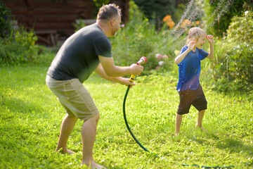Funny little boy with his father playing with garden hose in sunny backyard. Preschooler child...