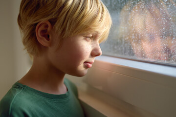 Portrait of preteen boy standing at the window. Child watching the rain outside.