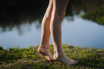 A barefoot girl, a teenage child walks along a lawn with green grass near the sea in the summer at sunset. Close-up photo of legs in nature.