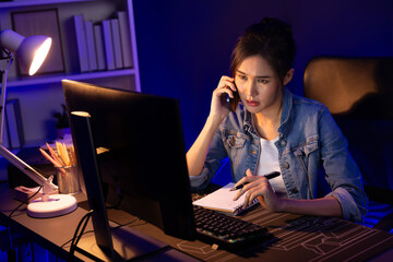 Young beautiful Asian creative woman calling on phone desk to customer or coworker explaining creator project's high profit while searching market analysis on pc at neon office at night. Stratagem.
