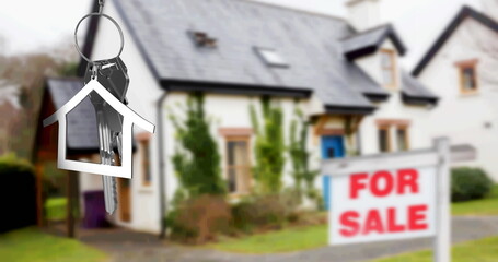 Image of silver house key fob and key, hanging in front of blurred house with for sale sign