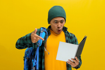 An excited young Asian student, dressed in a beanie hat and casual shirt, carrying a backpack, appears surprised while engrossed in reading a book against a yellow background