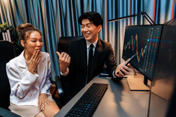 Asian businessman showing stock market application on mobile phone to partner pointing by pen, comparing financial digital investment database on computer screen at neon light at night time. Infobahn.