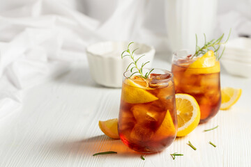 Iced tea or alcoholic cocktail with ice, rosemary and lemon slices on the white table.
